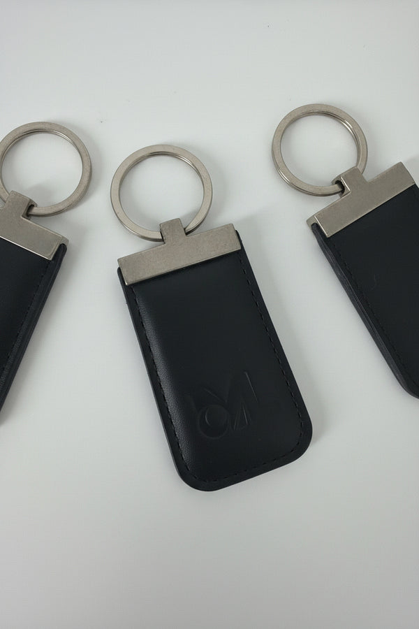A black leather key fob case with an embossed logo, attached to a silver metal split ring, shown on a plain white background.
