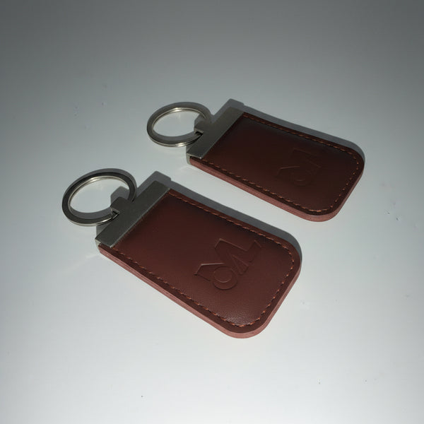 Two brown leather key fob covers with silver metal key rings and plates, featuring embossed initials on a light gray background.