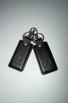 Two black leather key fob covers with silver metal keyrings and snap buttons, featuring an embossed logo on each cover, shown on a light gray background.