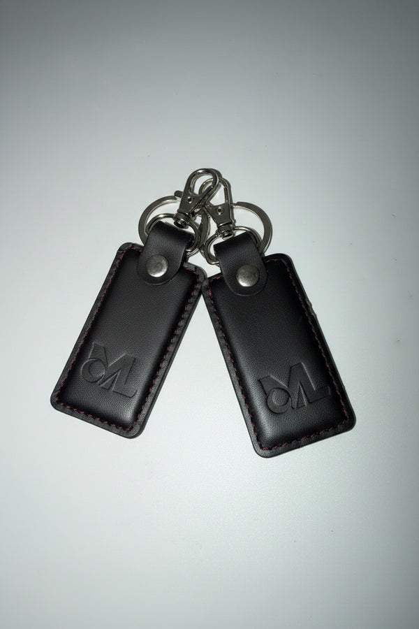 Two black leather key fob covers with silver metal keyrings and snap buttons, featuring an embossed logo on each cover, shown on a light gray background.